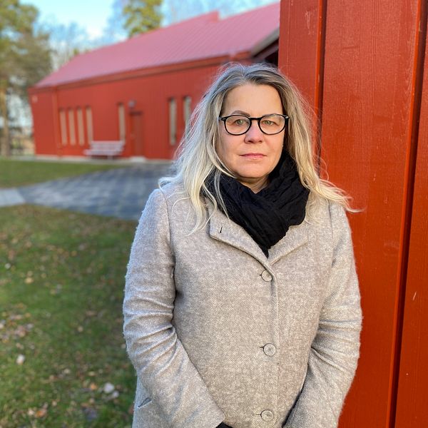Madeleine Karlsson, verksamhet F-6 i Motala, i en grå kappa och svart scarf står intill en röd vägg i en parkliknande miljö.