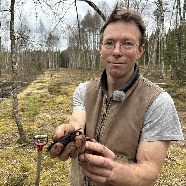 Skogsägaren Simon Jonegård håller i en bunt torv i en skogsmiljö, våtmark, med bäck och mossa.