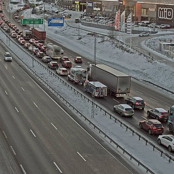 Kö av bilar och lastbilar på en snöbelagd motorväg med flera filer. Den övre delen av bilden visar ett trafikledningssystem och en kommersiell byggnad i bakgrunden.