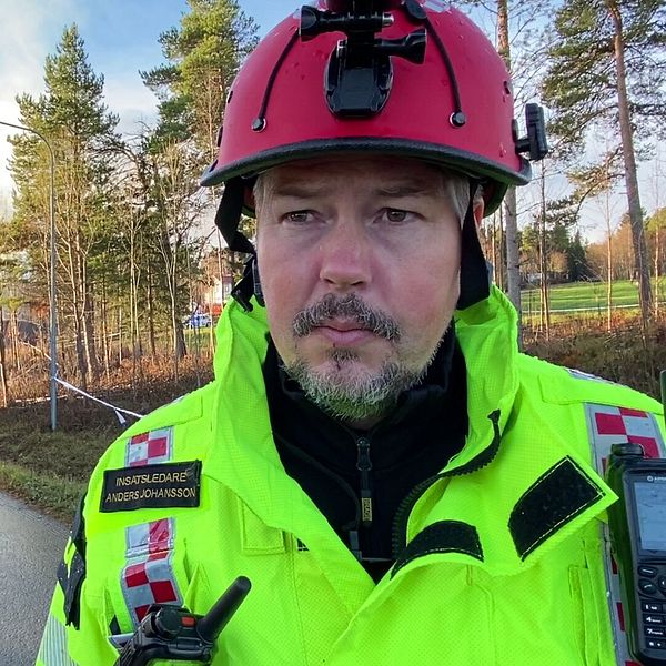 En brandman i en gul och svart skyddsutrustning står framför en räddningsinsats på en väg. I bakgrunden syns en brandbil med blåljus, samt flera andra fordon på platsen. Träden i området ger en naturnära känsla, och det är en klar himmel. Situationen tyder på en aktiv räddningsoperation.