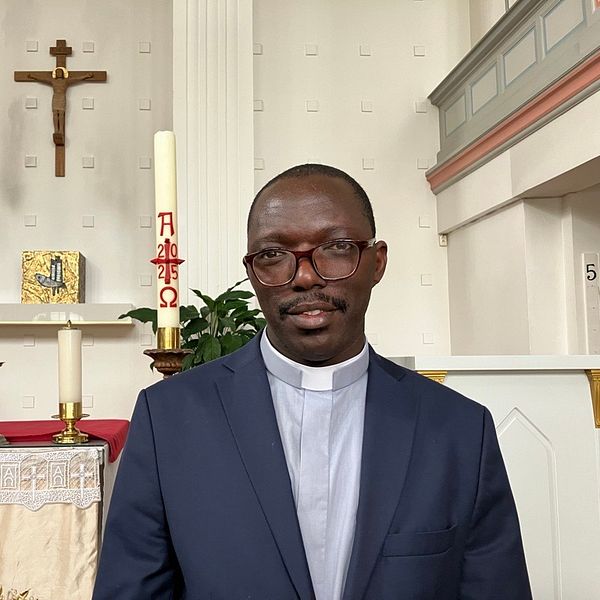 Präst i kavaj står framför altaret i en kyrka med kors och ljus i bakgrunden.