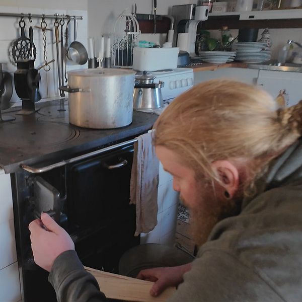 Mio Fessé sitter på knä och arbetar med en gammaldags vedspis i ett kök. Spisen är svart och av metall, och en stor kastrull står på plattan.
