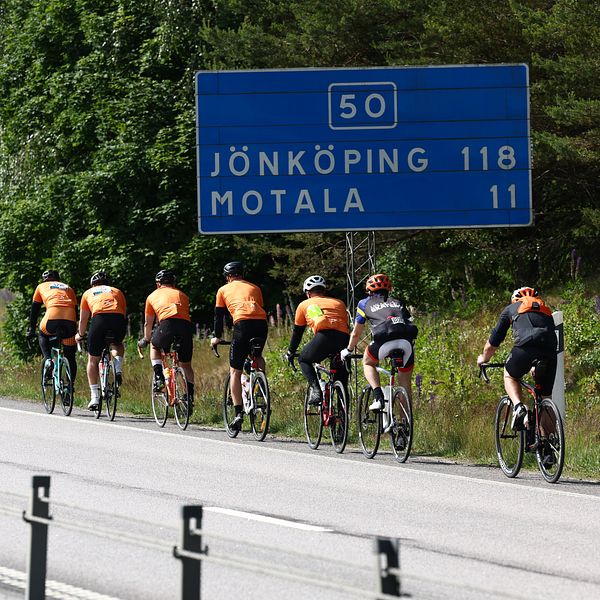 En grupp cyklister i orangea tröjor cyklar längs en väg. Bakom dem syns en vägskylt som visar avståndet till Jönköping (118 km) och Motala (11 km). I bakgrunden är en grönskande skog och en bilväg synlig.