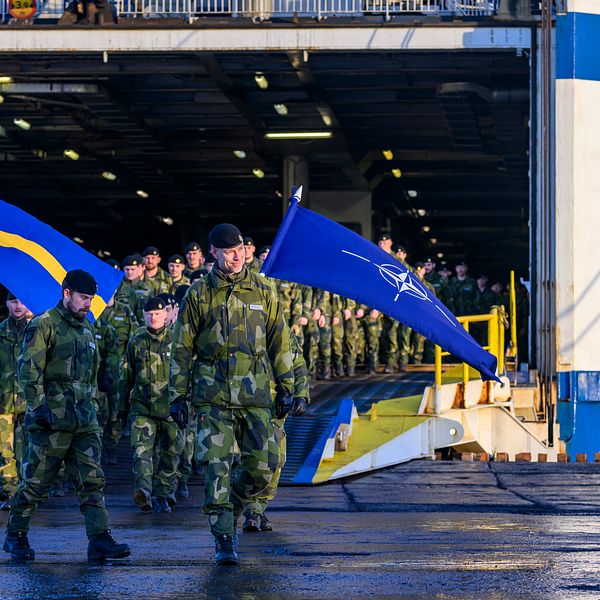 Svenska soldater marcherar ut från ett militärt transportfartyg, bärandes flaggor med svenska och NATO:s symboler. I bakgrunden syns flera soldater i uniform som följer efter i ledet.