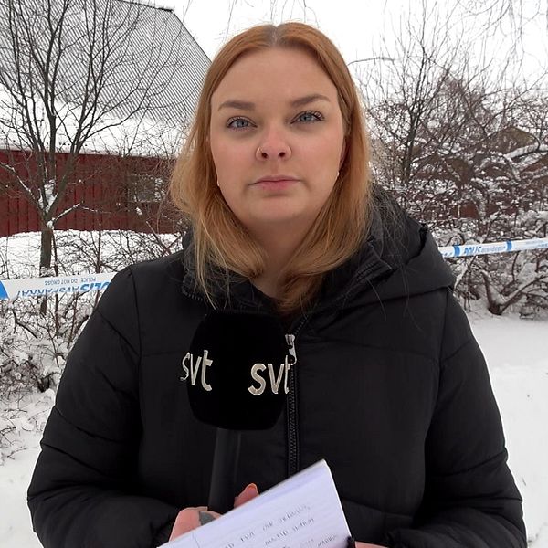 En reporter från SVT står utomhus med en mikrofon i handen, bredvid ett område som har avspärrats av polis. I bakgrunden syns en snötäckt björk och några byggnader.