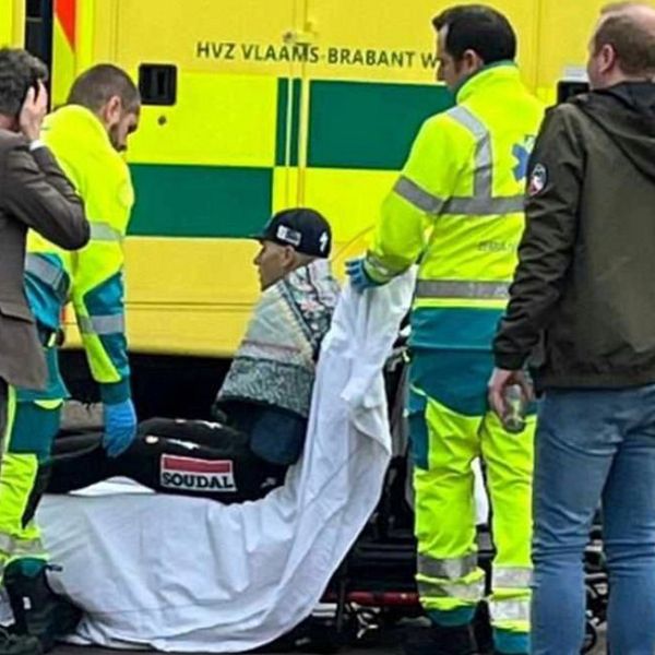 En grupp räddningspersonal assisterar en skadad person på en bår, täckt med en vit filt. Två av räddningsarbetarna har på sig reflexkläder och en tredje bär en jacka med logotyp. I bakgrunden syns en ambulans med tydliga märkningar. Enligt situationen förefaller det vara ett akut ärende, med flera personer närvarande, inklusive en polis.