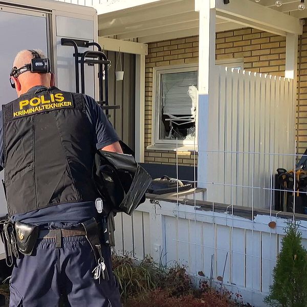 En polis på en brottsplats arbetar vid en bil. I bakgrunden syns en bostad med ett trasigt fönster.