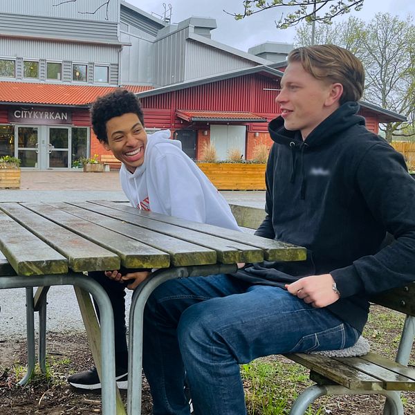 Tre personer sitter vid ett utomhusbord framför en byggnad. En av dem skrattar.
