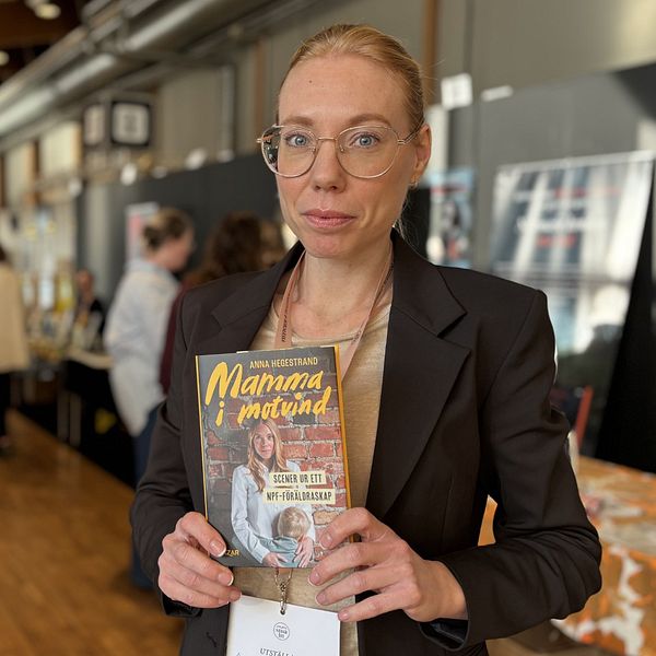 En person håller upp en bok med titeln ”Mamma i mittend” på en bokmässa där människor bläddrar och samtalar vid olika bokstånd i bakgrunden.