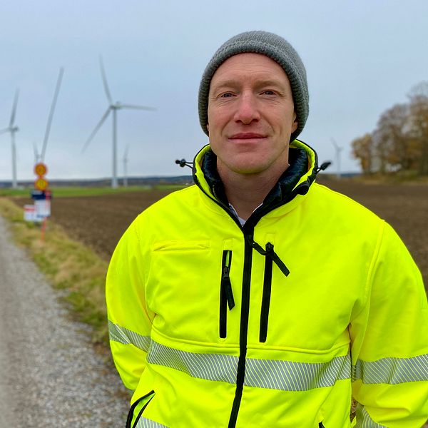 Man i gula varselkläder vid en väg. Vindkraftverk i bakgrunden