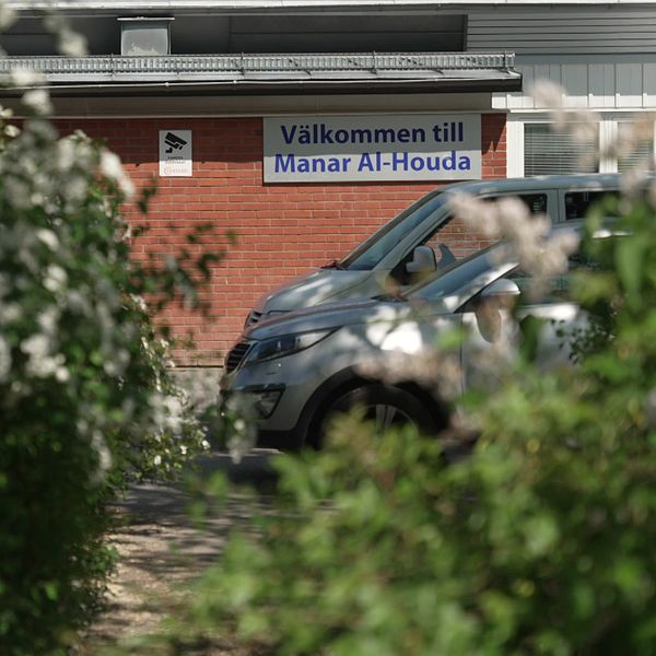 Buskage i förgrunden. Längre bort står bilar parkerade och skolans skylt,