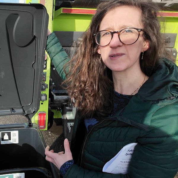 Person i grön jacka visar öppna återvinningskärl framför en lastbil.