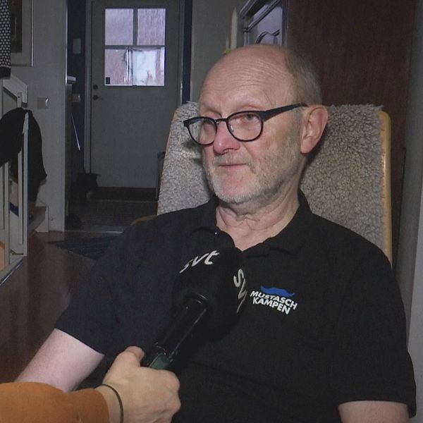 Person i fåtölj intervjuas inomhus, mikrofon riktad mot personen.