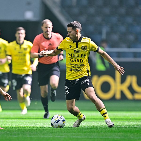 Fotbollsmatch mellan lag i gula och svarta tröjor, spelare tävlar om bollen på planen.