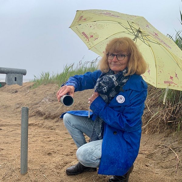 Eva Hallberg sitter på knä under ett paraply framför en vägbom på en strand som sågats av.