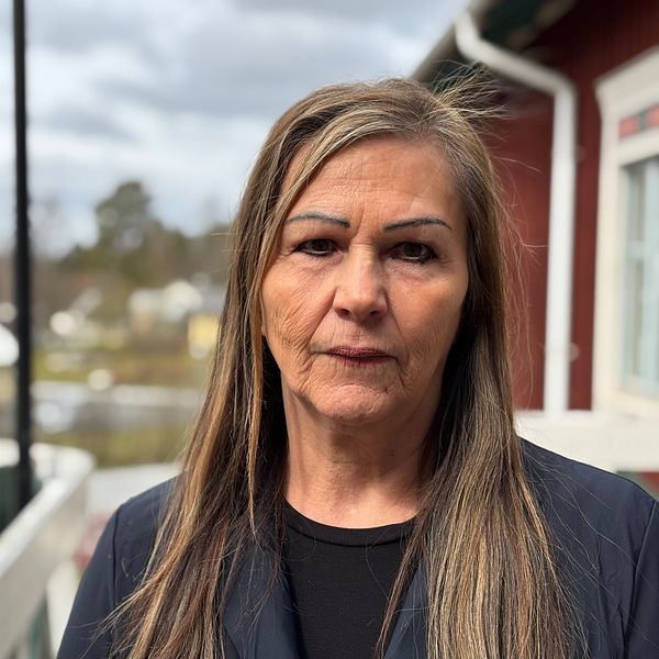 Kvinna med långt hår står utomhus vid ett hus, bostadsområde syns i bakgrunden.