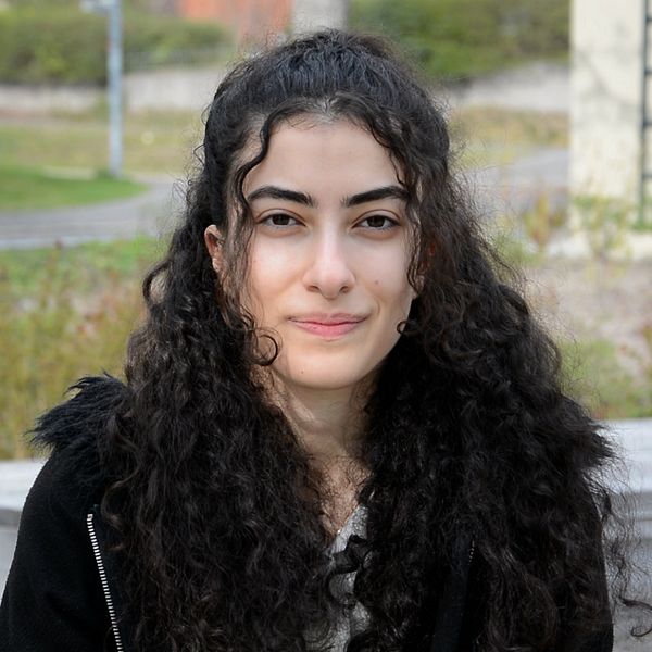 Person med långt lockigt hår sitter utomhus på en bänk, grönska i bakgrunden.