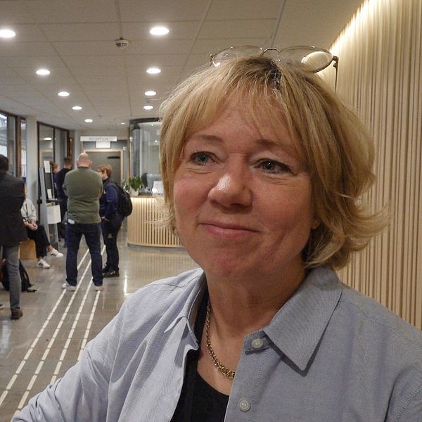 En kvinna står vid ett bord i en modern och ljus lokal, med en avslappnad atmosfär. I bakgrunden syns personer som väntar i en reception. Inredningen är stilren med trädetaljer och växter som bidrar till en vänlig miljö.