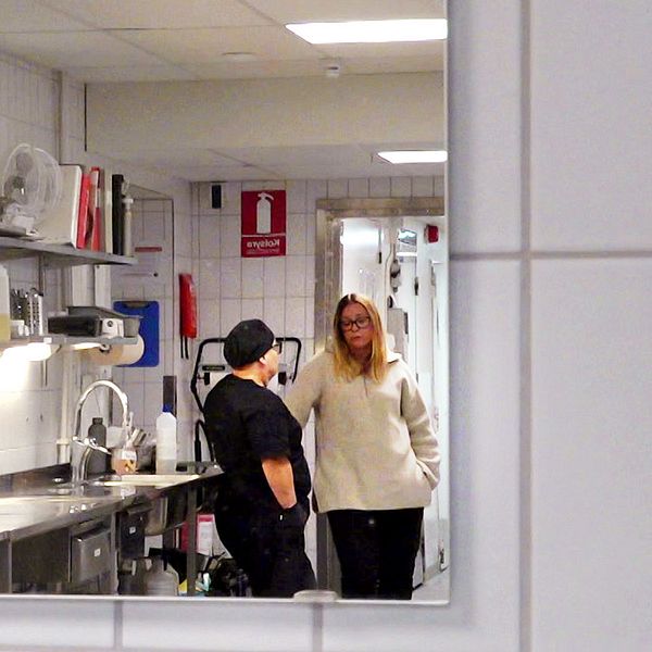 Två personer samtalar i en lukas köksmiljö, med en rad köksredskap och förvaringsbehållare bakom dem. Den ena personen står vid en disk, medan den andra står i närheten och lyssnar. Rummet är upplyst och ser välorganiserat ut.