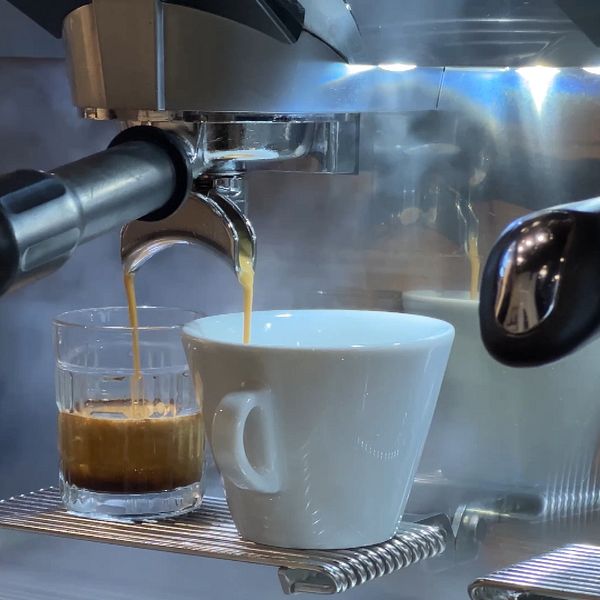Espresso maskin som brygger en espresso, med en liten glasbehållare för espresso och en vit kopp under utloppsröret. Dämpad ånga stiger från maskinen.