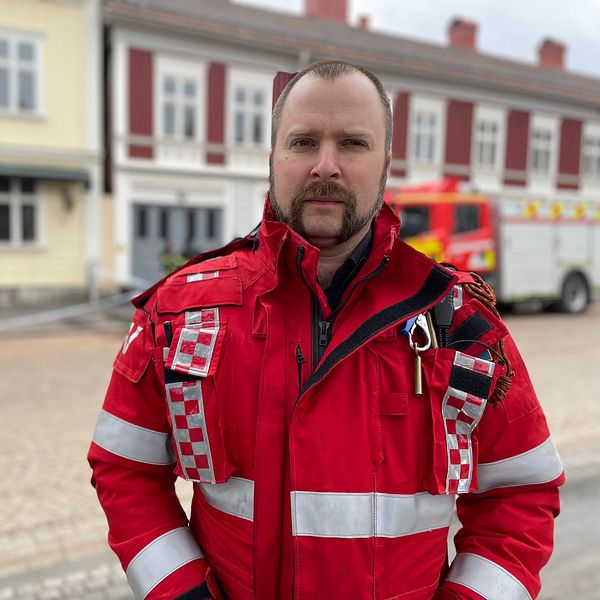Brandman i röd uniform framför brandbil och byggnader.