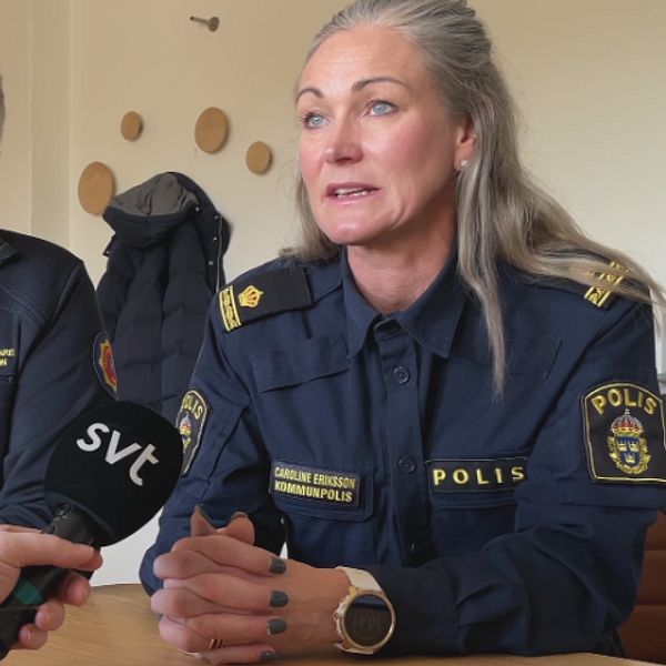 En intervju sker mellan två personer, där en person bär en polisuniform med märket ”POLIS” och den andra har en jacka med texten ”SÄKERHETSANSVARIG.” En mikrofon hålls i bild, som visar att det handlar om en medieintervju. Bakgrunden har en enkel inredning och en jacka hänger på väggen.