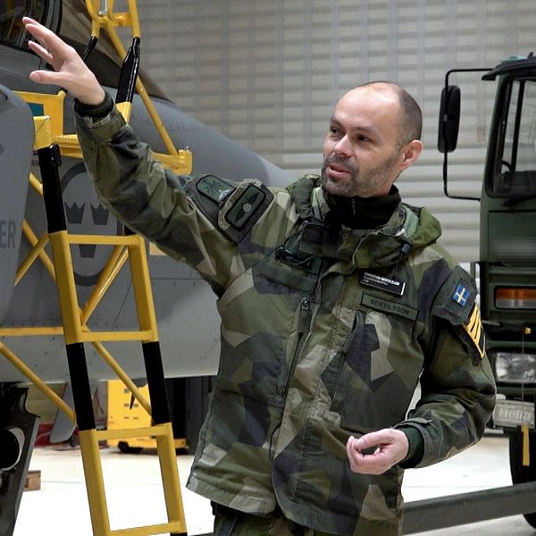 En person i militäruniform pekar mot en del av ett flygplan inuti en hangar, med en stege placerad intill. I bakgrunden syns en militär lastbil och andra detaljer från hangaren.