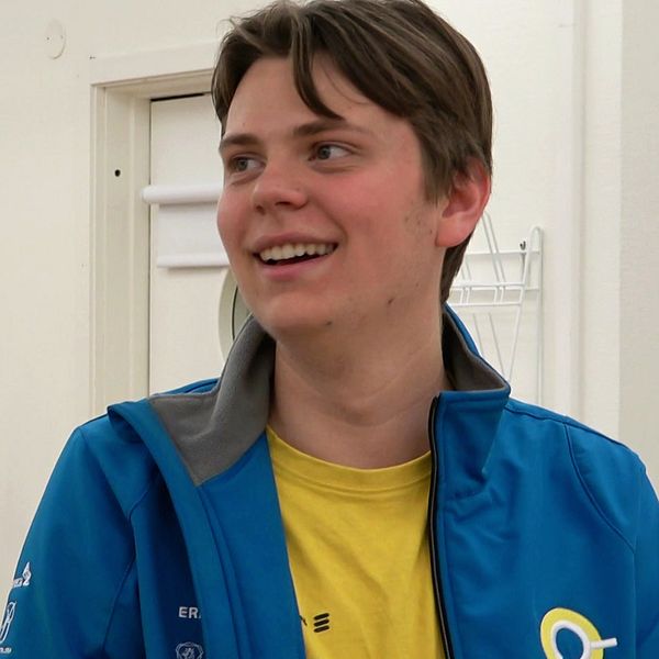 Person i blå jacka med gul tröja står i ett laboratorium, vita rockar hänger på väggen.