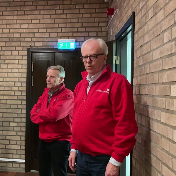 Fyra personer i röda jackor står i en inomhusmiljö med tegelväggar. Belysning kommer från taklampor och en dörr syns till vänster i bilden.