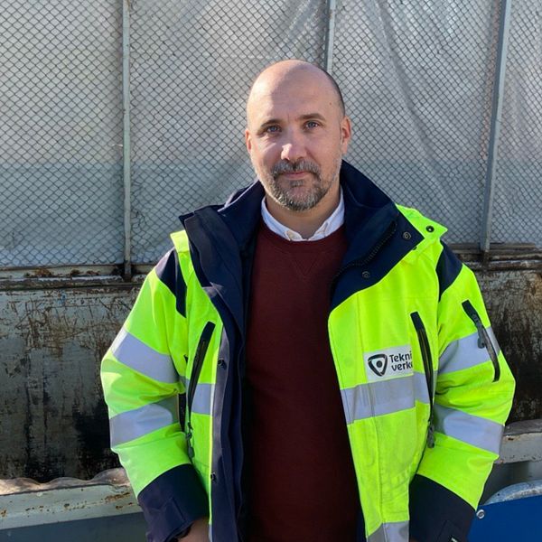 Bildens vänstra del visar en skylt som anger ”Energiåtervinning,” med en tydlig blå himmel i bakgrunden. Till höger står Johan Borg klädd i en reflekterande jacka i gul och blå. Bakgrunden är en container.