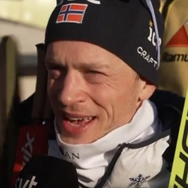En skidåkare i tävlingskläder med norska flaggan synlig på mössan står i vad som verkar vara en tävlingsmiljö, med människor och färgglada banderoller diffust i bakgrunden. Skidåkaren har en stav och en mikrofon synlig framför sig, vilket tyder på en intervju eller en kommentar till media.