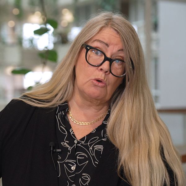 Annika Wallenskog med långt, blont hår gestikulerar med sin hand i en inomhusmiljö. Bakgrunden visar en modern byggnad med stora fönster och växtlighet, vilket skapar en ljus och öppen atmosfär. Personen är klädd i en svart tröja med mönster.