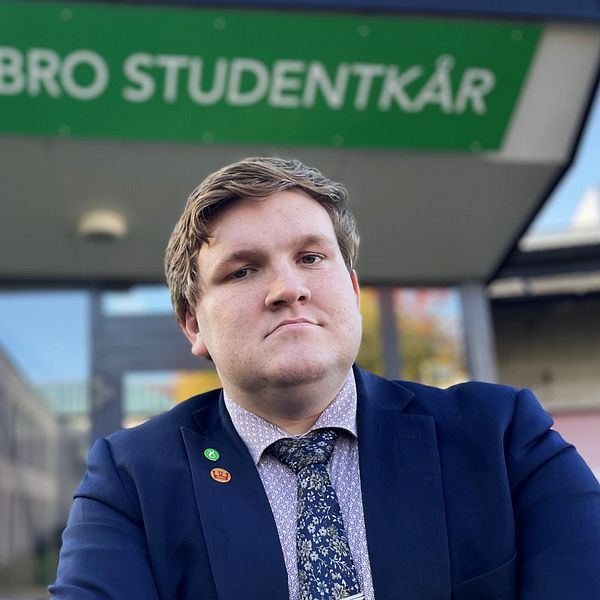 En person i kostym står framför Örebro Studentkårs lokaler. Byggnaden har en tydlig grön skylt med namnet.