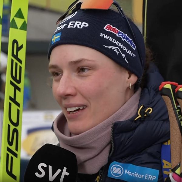 En idrottare med skidor från Fischer står vid en reporter med mikrofon i en spännande tävlingsmiljö, med publiken synlig i bakgrunden.