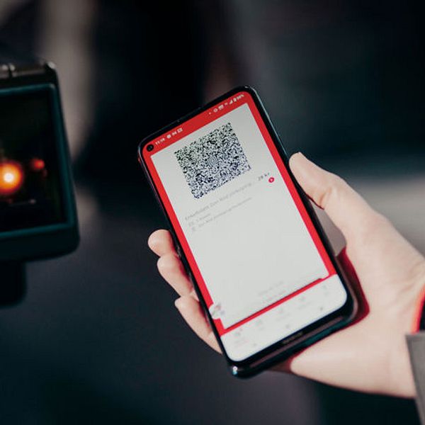 En person håller upp en smartphone med en QR-kod mot en QR-kodsläsare. Skärmen på telefonen visar en app med QR-koden och tillhörande information. Bakgrunden är suddig och fokuserar på interaktionen mellan telefonen och läsaren.