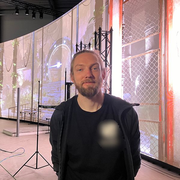 En man står framför en stor,  rundad bildskärm med projektioner av en grönskande, drömlik scen. Rummet är inrett med olika strukturer som stödjer ljus- och ljudutrustning, och det finns ledningar på golvet som tyder på en pågående produktion eller inspelning.