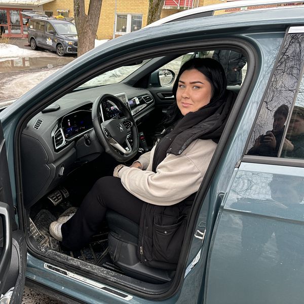 Person sitter i förarsätet på en bil med öppen dörr utomhus.