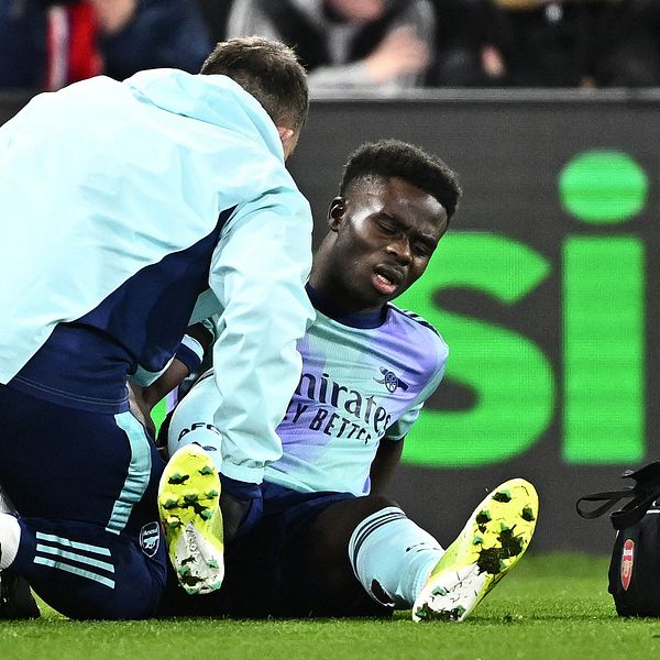 En fotbollscoach assisterar en spelare som sitter på marken efter att ha blivit skadad under en match. Spelaren bär en ljusblå tröja med klubbens logotyp och ser ut att få medicinsk hjälp.