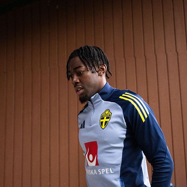 En fotbollsspelare i träningskläder promenerar vid trävägg.