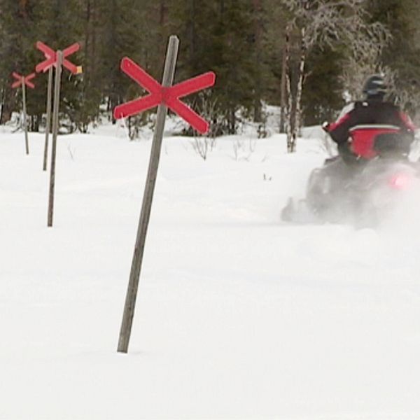 Till vänster står en person klädd i en mörk jacka med reflexer, i en skogsmiljö. Till höger visas en snötäckta landskap med flera röda kryssmarker som står upprätt i snön, och en snöskoter som färdas förbi.
