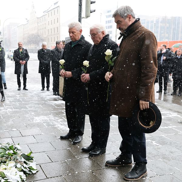 Människor deltar i en minnesceremoni vid en plats täckt med blommor under en snöig dag. Tre män håller vita rosor framför dem medan andra deltagare och åskådare står i bakgrunden.