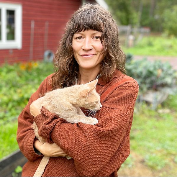 kvinna håller i katt, står framför odlingar vid rött trähus