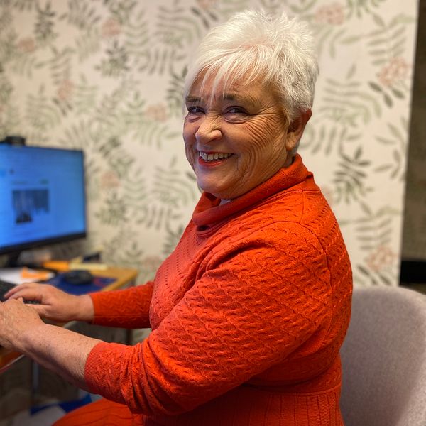 En kvinna, Helen Andersson, sitter i en röd klänning vid sin dator  som står på ett skrivbord.