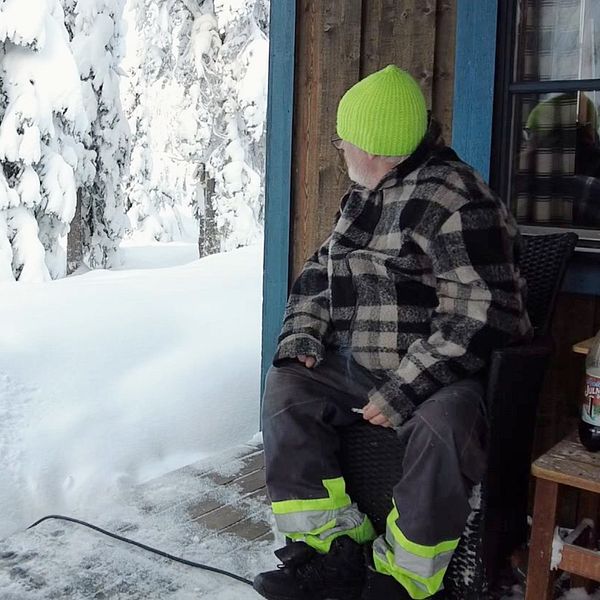 En man i neongrön mössa sitter på en altan till en stuga i Sälen och tittar ut på ett tjockt snötäcke.