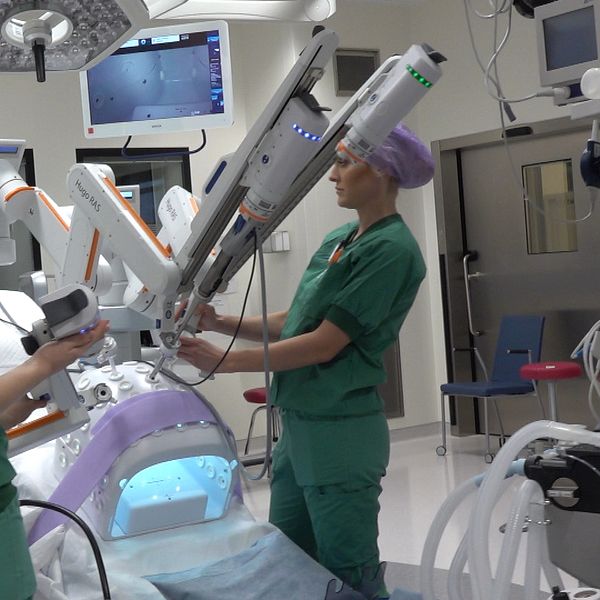 Två sjukvårdspersonal klädda i gröna operationskläder står vid en avancerad kirurgisk robot i ett operationsrum. Den ena personen håller i en kontroll och fokuserar på robotens instrument, medan den andra justerar maskinens position. I bakgrunden syns flera skärmar som visar olika medicinska data och bilder. Rummet är modernt och välutrustat för kirurgiska ingrepp.