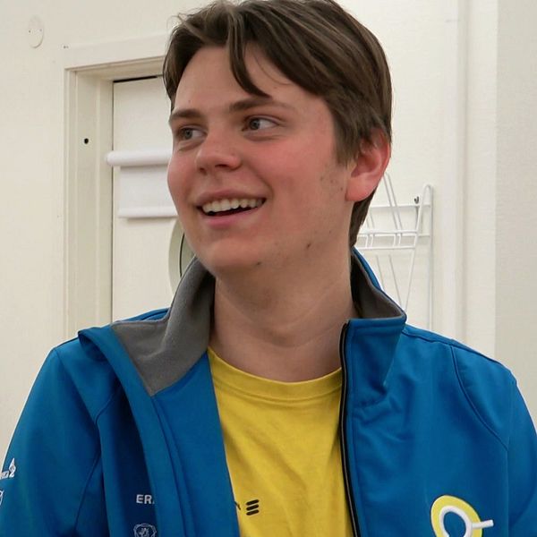 Person i blå jacka och gul tröja står i ett rum med laboratorierockar på väggen.