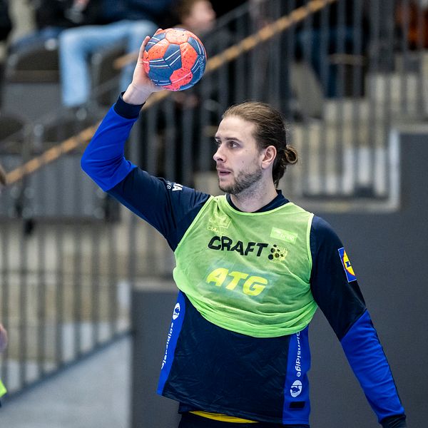 En handbollsspelare i en grön träningströja förbereder sig för att kasta en handboll, medan en annan spelare står i bakgrunden. Arenan har åskådare i bakgrunden och ger en känsla av en aktiv sportmiljö.