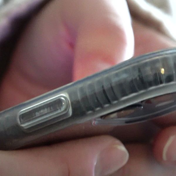 En hand håller i en smartphone, med fokus på en del av telefonens kant och en knapp. Fingrarna är aktiva och skärmen avger ett svagt ljus. Bakgrunden är suddig och antyder en inomhusmiljö.