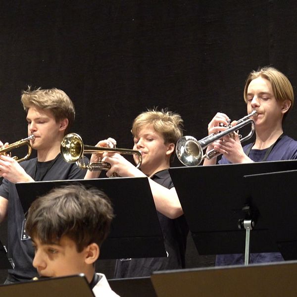 FEM UNGDOMAR STÅR OCH SPELAR TRUMPET I ETT STORBAND.