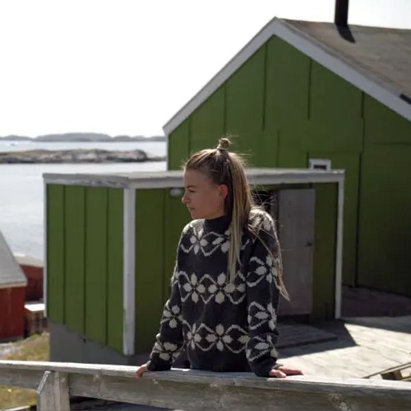 Kvinna i mönstrad tröja blickar ut över traditionella hus vid kusten i en nordlig miljö.